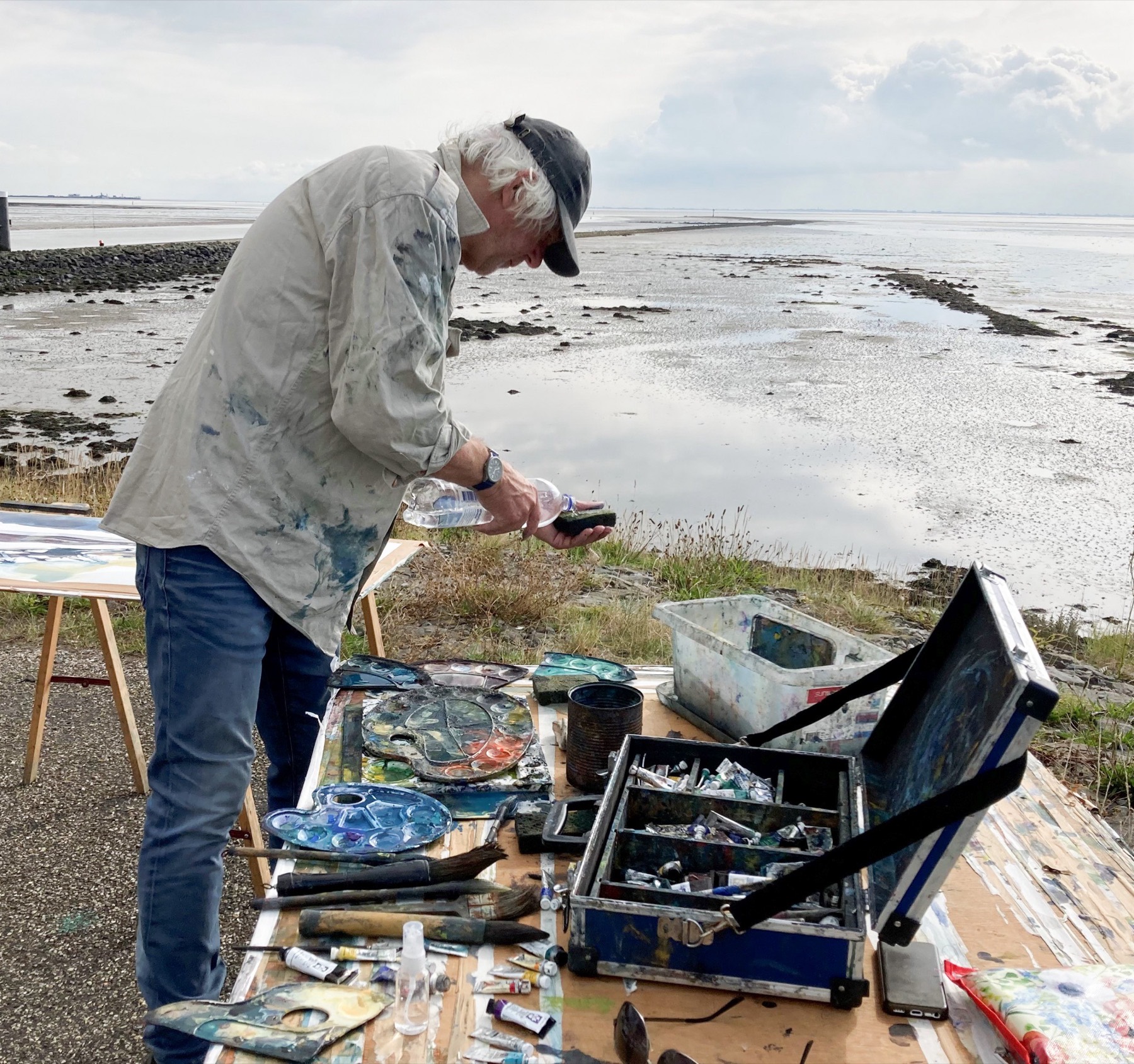 Johan Meeske aan het werk op de kwelder: hij giet water op een spons, voor hem een tafel vol paletten, verftubes en kwasten, achter hem het wad bij laag water.